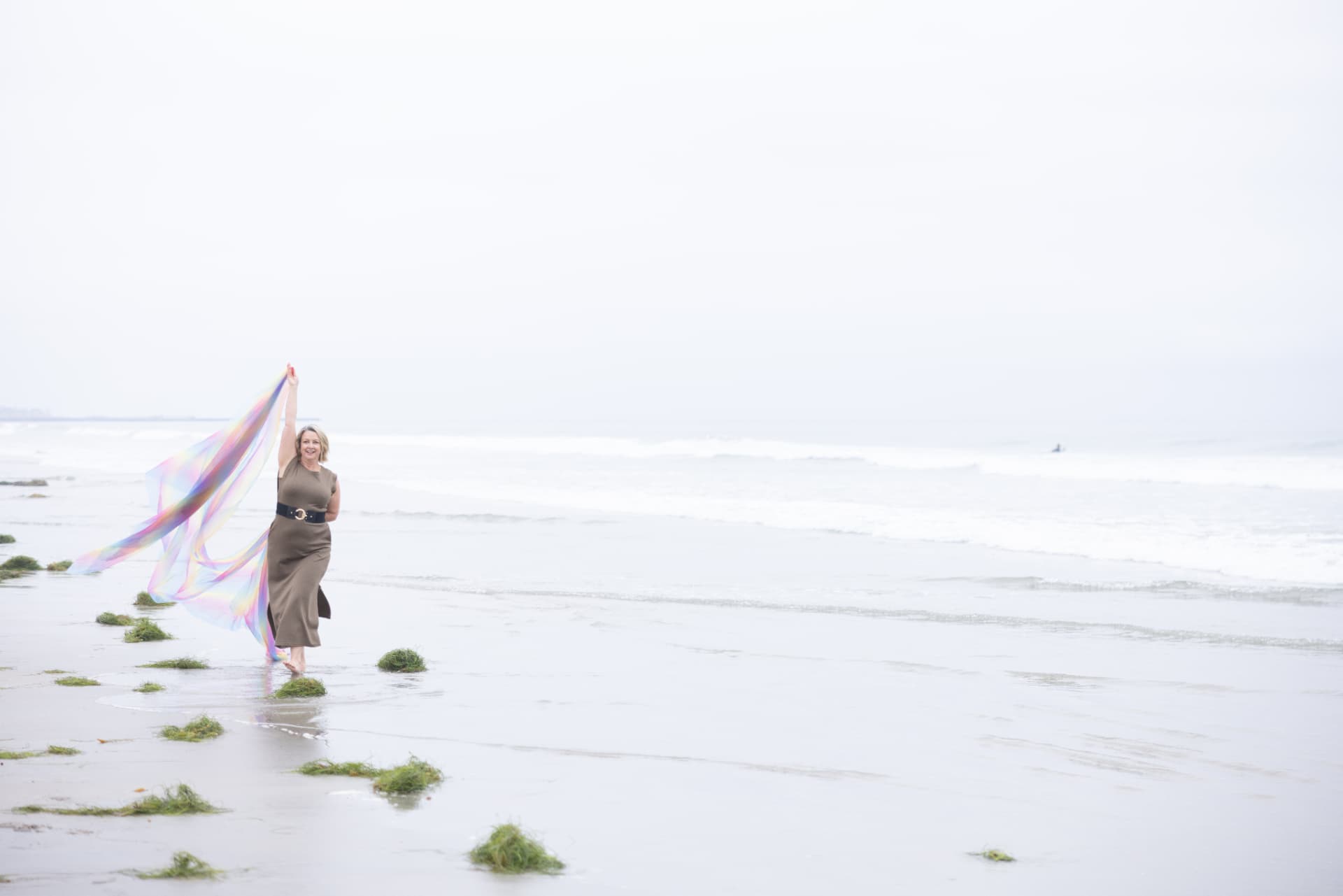 A woman walking barefoot along the shoreline holding an iridescent scarf above her head