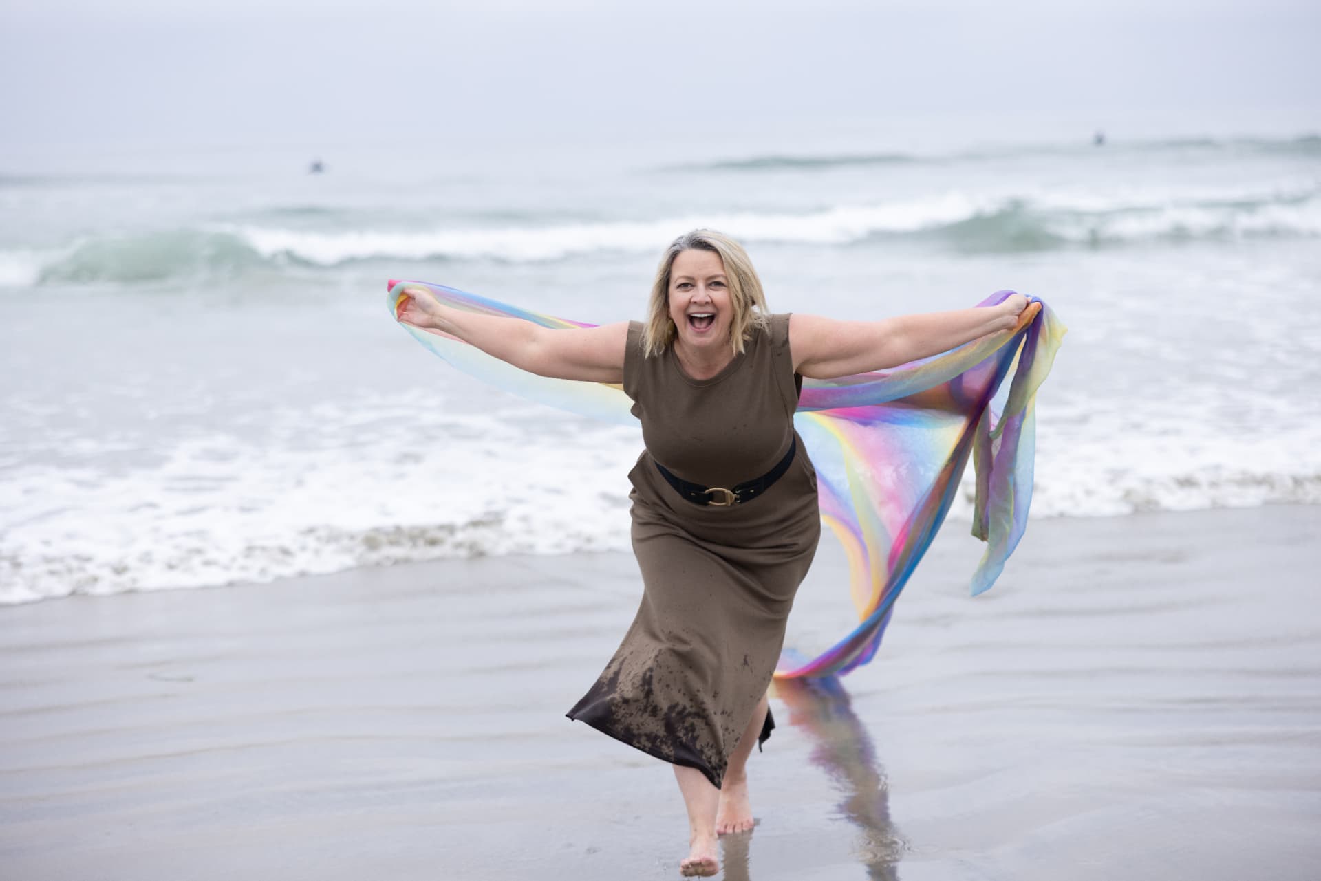 D LeD laughing at the shoreline with a rainbow scarf flowing behind her